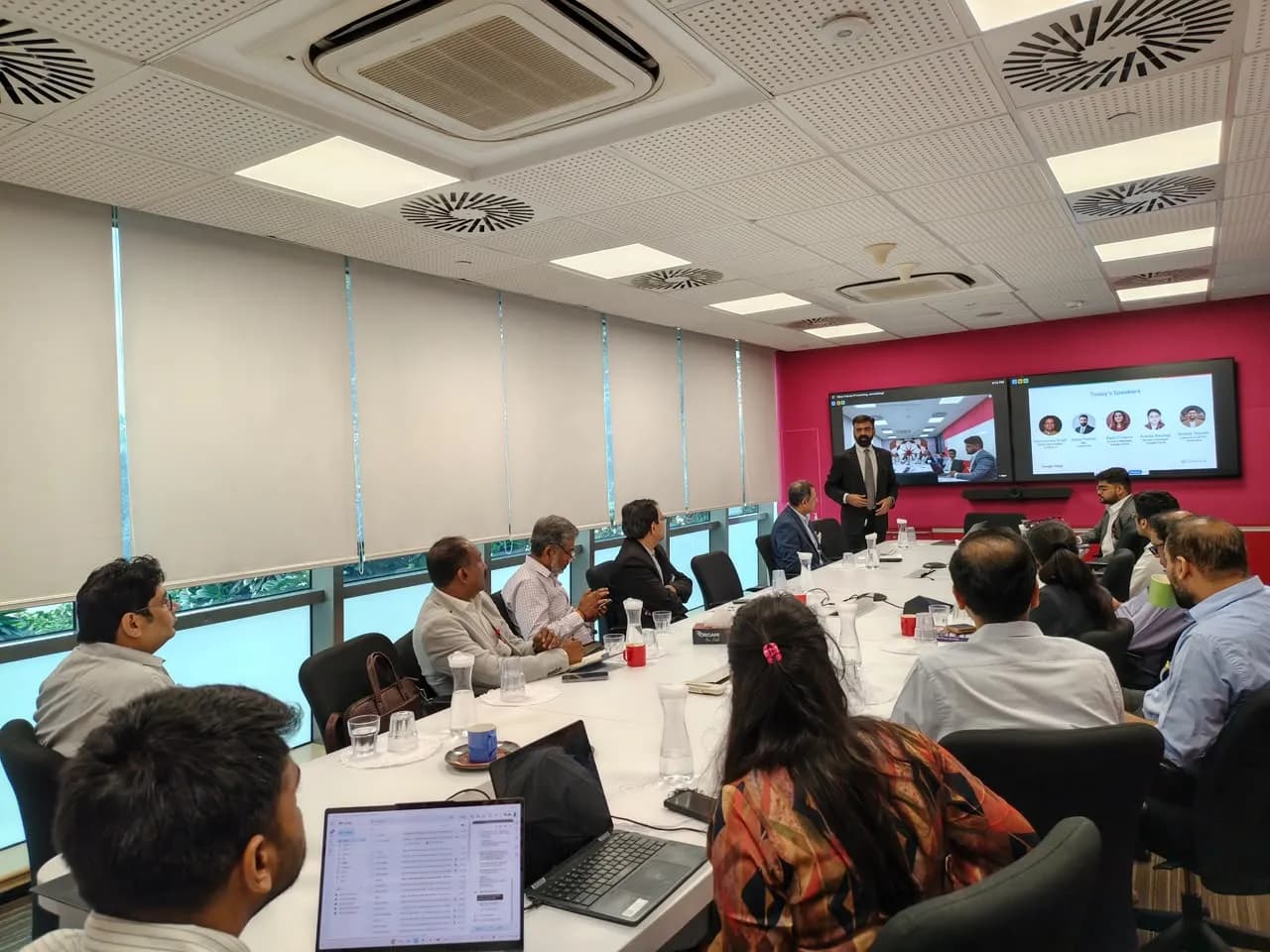 Business leaders seated along the boardroom table facing two screens at the far end
